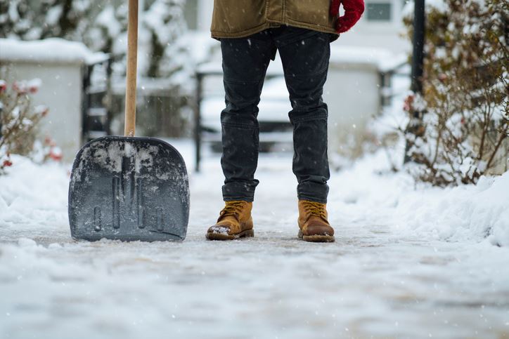 Two lower legs in boots with a snow shovel with a freshly cleared snowy driveway in the background